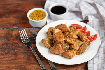 Fried and stewed chicken breast in soy-honey sauce. In the background mustard seeds. On a plate are slices of tomato and razmarin. On a wooden background.