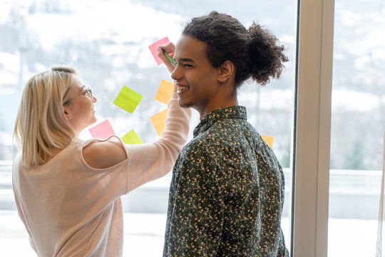 Positive Young People Laughing While Working Together During Brainstorming And Standing Behind Glass Wall With Sticky Colorful Papers.Cheerful Students Learning Words From Stickers