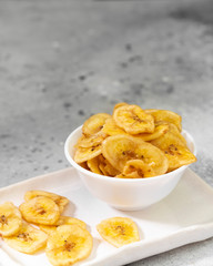 Dried bananas in a white ceramic bowl on a gray kitchen table. Banana chips. Vegetarian snack for proper nutrition