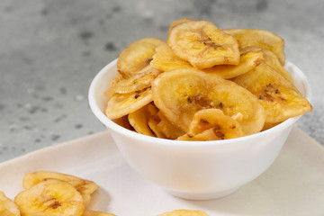Dried bananas in a white ceramic bowl on a gray kitchen table. Banana chips. Vegetarian snack for proper nutrition