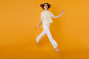 Fototapeta na wymiar Full-length portrait of glad girl in yellow t-shirt and white pants running on orange background. Studio shot of wonderful caucasian woman dancing with pleasure.