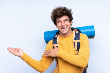 Young mountaineer man with a big backpack over isolated blue background extending hands to the side for inviting to come