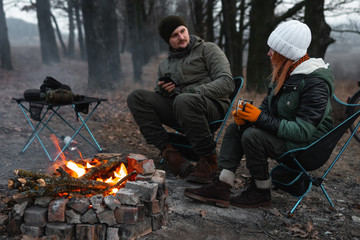 A couple of people are sitting on light chairs by the fire. Cold autumn evening in the forest. active lifestyle, travel, vacation. campfire. have a conversation over a cup of tea, dialogue