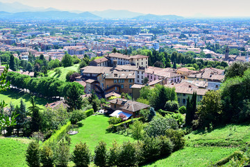 Fototapeta premium Bergamo, Italy - Panorama, mountain view of the city with tiled roof houses, green fields with trees, in the summer afternoon. Описание (на английском языке)127/