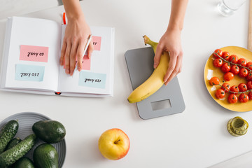 Top view of girl putting banana on scales while writing calories in notebook, calorie counting diet - obrazy, fototapety, plakaty