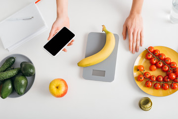 Top view of girl holding smartphone while weighing banana on kitchen table, calorie counting diet