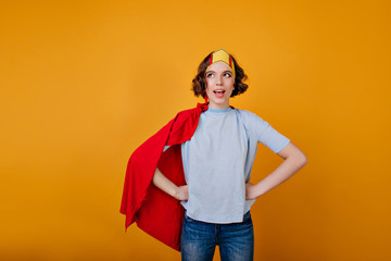 Enthusiastic girl with curly hairstyle posing in red cloak. Indoor photo of ecstatic female superhero in toy crown.