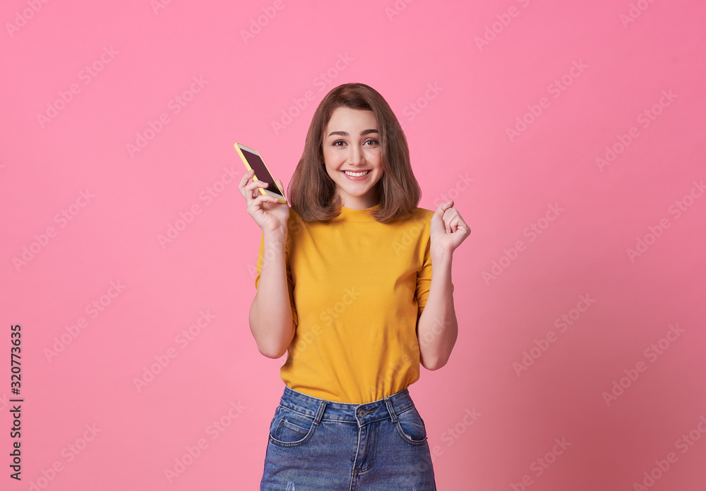 Wall mural Portrait of happy young woman holding mobile phone isolated over pink background.