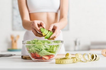 Cropped view of slim woman cooking salad with fresh vegetables and avocado near measuring tape on...