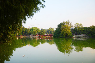 Fototapeta premium Morning time at the Huc Bridge—a wooden, red-lacquered piece of olden Vietnamese architecture that provides a spectacular view of the lake and its surroundings at Hoan Kiem Lake.