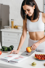 Selective focus of beautiful smiling sportswoman writing calories while weighing apple on kitchen...