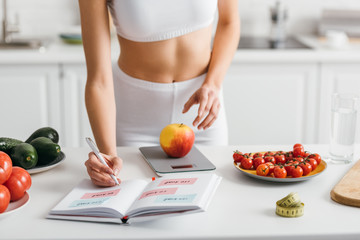 Cropped view of fit sportswoman writing calories while weighing apple on kitchen table, calorie counting diet
