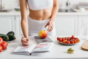 Poster Cropped view of fit sportswoman writing calories while weighing apple on kitchen table, calorie counting diet © LIGHTFIELD STUDIOS