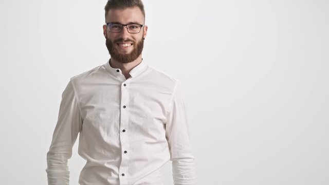 A positive young bearded man wearing a white shirt puts on glasses isolated over white wall background