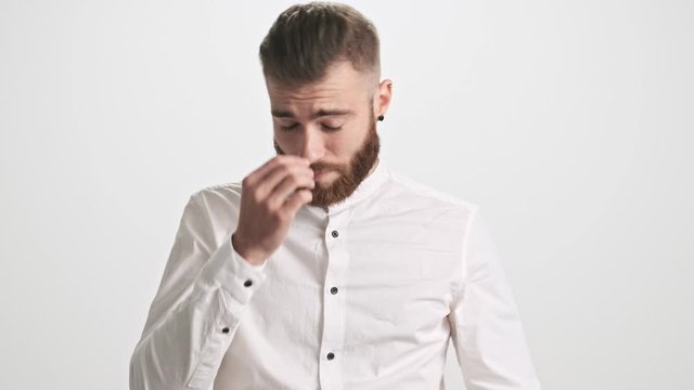 A tired young bearded man takes off his glasses and touches his bridge of nose and undo the top button of his shirt isolated over white wall background