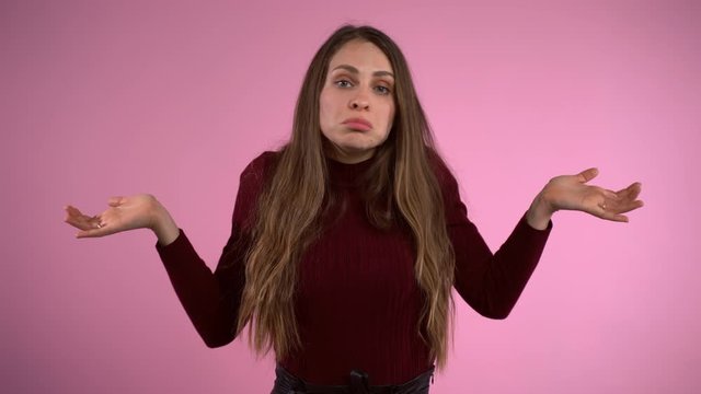 Portrait of pretty young long hair woman feeling perplexity and misunderstanding emotions posing isolated over studio pink background. Displeased woman throwing hands wide and shaking shoulders