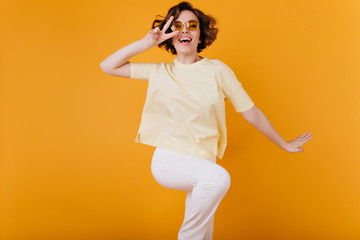 Fototapeta na wymiar Winsome short-haired girl in white attire dancing on orange background. Studio photo of gorgeous female model fooling around and laughing.