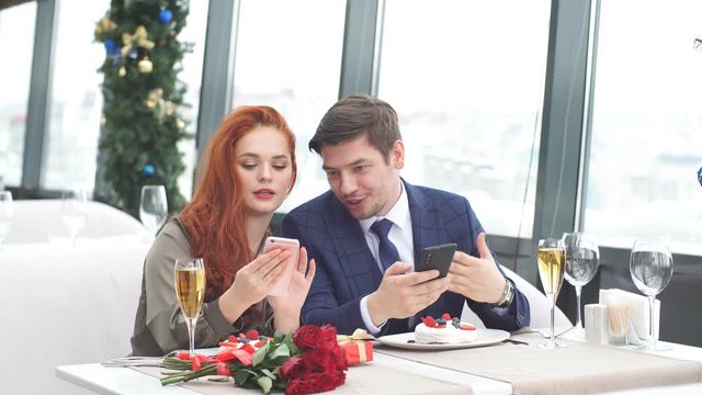Young European couple on a date in restaurant. Red-haired girl shows a guy pictures on his smartphone.