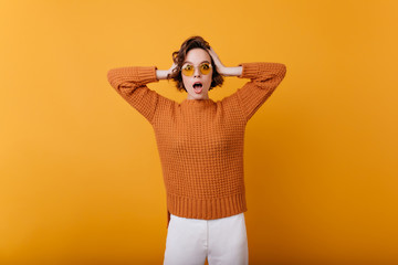 Charming woman with shocked face epxression touching her head. Studio shot of emotional european girl in trendy woolen sweater isolated on orange background.