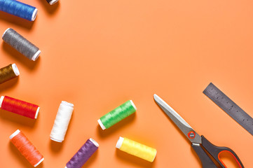 Scattered sewing accessories metal ruler, scissors and spools with colorful threads on orange tailors desk in workshop. Space for text. Top view