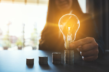 Businesswoman holding and putting lightbulb on coins stack on table for saving energy and money...