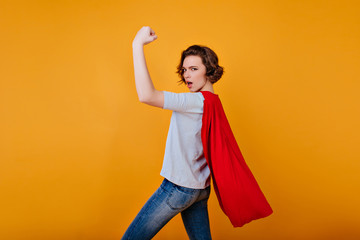 Strong girl in denim pants and blue t-shirt posing with hand up. Funny female model in red cloak...