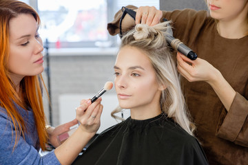 Women redhear make-up artist and hairdresser do hairstyle and stylish makeup for beautiful young blonde girl before start of fashion show. Colorful makeup design beauty and art salon concept