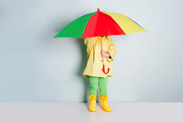 little child in rubber boots, yellow raincoat holding colored umbrella 
