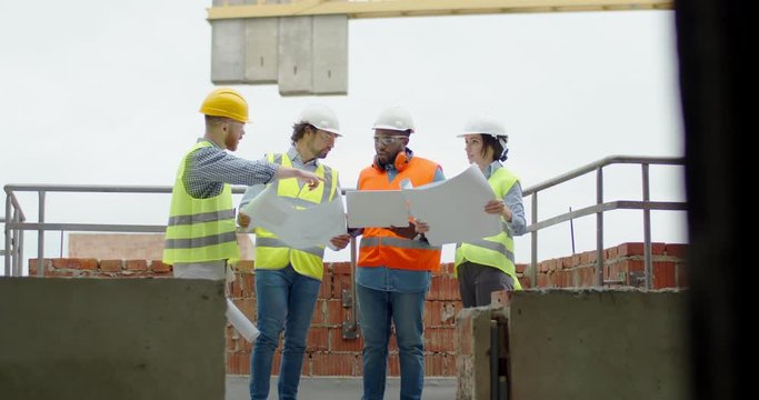 Caucasian woman architect and three mixed-races men builders with drafts in hands standing at the building site and talking about work.