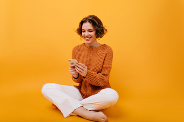 Smiling girl in casual outfit sitting with legs crossed and holding phone. Studio shot of gorgeous...