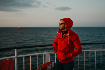 A stylish man with stubble in a red jacket in the hood and in the sunglasses puts on a hood on his...