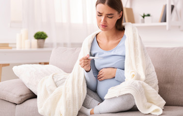 Unhappy pregnant lady having fever feeling bad sitting on sofa