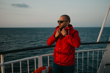 A bald man with stubble in a red jacket in the sunglasses fastens jacket aboard a ship looking at the sunset.