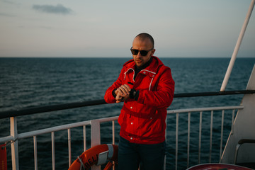Obraz premium A bald man with stubble in a red jacket in the sunglasses checks the time on a watch aboard a ship