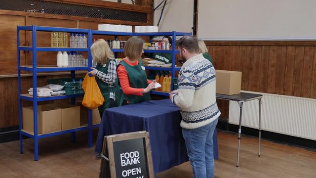 Man collects two bags of free food from a food bank. Volunteers work hard in the background to sort out the food donations. 
