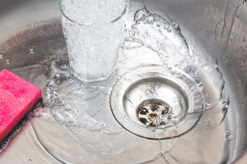 A pink dishwasher sponge lies in a metal sink. Nearby is a glass with water