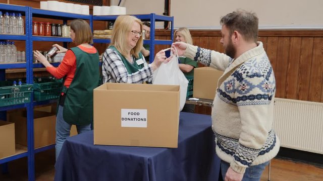 Man donating a bag of food to a foodbank. Volunteers work hard in the background to sort out the food donations. 