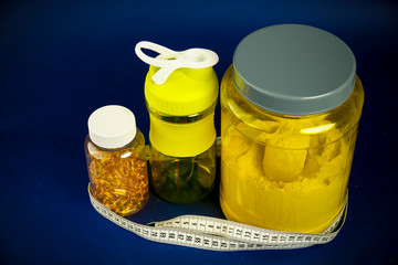 Protein in a yellow jar, plastic shaker, green dumbbells and an omega 3 jar on a blue background