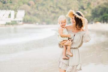 Fototapeta na wymiar Happy woman mom in a hat and her baby in her arms on a tropical beach. Summer vacation concept and family look.