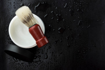 Top view of jar of foam for shaving and brush with wooden handle, black surface with water...