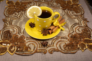 A cup of tea with lemon stands on a beautiful oriental napkin. On a saucer are sticks of cinnamon and star anise..