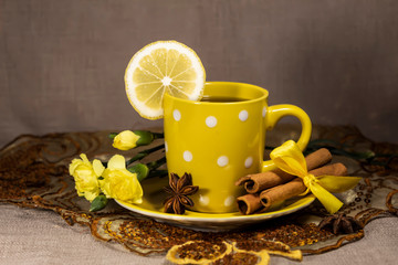 A cup of tea with lemon is on a napkin. On a saucer lies star anise, cinnamon sticks and yellow clove flowers.