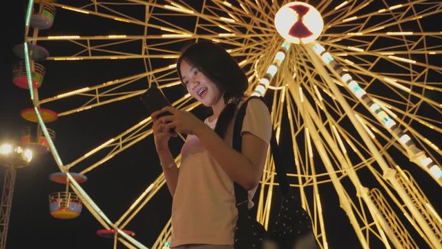Happy beauty Asian woman look at smart phone chatting with friend in Amusement park background with with ferris wheel ride background. People lifestyle and Entertainment. Travel and holiday vacation
