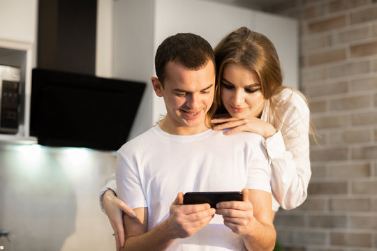 Close Up Of A Couple In Love Using A Cellphone Together In The Kitchen. Woman With Long Hair Hugging A Man From Behind