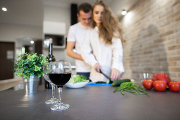 Portrait of romantic couple in the kitchen at home. Blonde woman with long hair slices cucumber. A man hugging a woman from the side. A glasses with red wine in the foreground