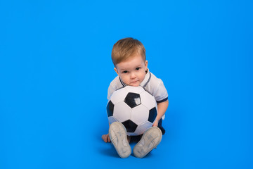 Boy in a football uniform sits on a blue background with a soccer ball and looks forward.