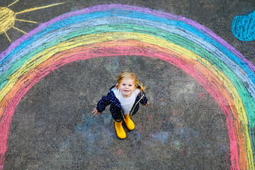 Happy little toddler girl in rubber boots with rainbow sun and clouds with rain painted with colorful chalks on ground or asphalt in summer. Cute child having fun. creative leisure