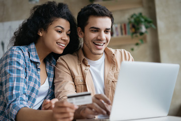 Portrait of beautiful multiracial couple shopping online, using laptop computer.  Happy friends ordering food on website. Smiling African America woman holding credit card, looking at digital screen 