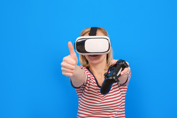 Young girl in white trousers and striped sweater is playing virtual reality with helmet VR and gamepad on a blue background. Innovative technologies and concept of education and entertainment.