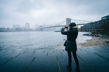 Tourist woman with warm clothes takinga photo with the Brooklyn Bridge on the background in DUMBO...